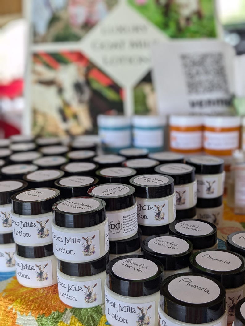 May include: A display of small jars of goat milk lotion with black lids and white labels. The labels feature a goat illustration and the words "Goat Milk Lotion." Various scents are labeled, including "Peaceful Lavender" and "Plumeria."