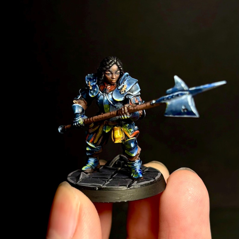 May include: A blue and gold painted miniature figurine of a female warrior wearing armor and holding a spear. The figurine is standing on a black base.