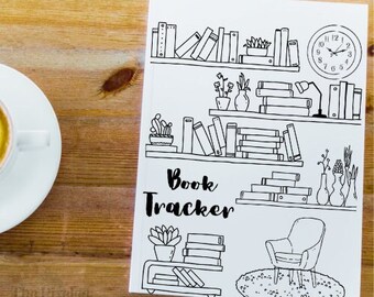 May include: A white "Book Tracker" journal with black line art of bookshelves, a clock, and a reading chair. The journal is on a wooden surface, with a cup of coffee in the upper left corner.