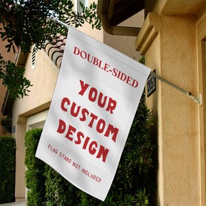 May include: A white, double-sided flag with red text that reads "DOUBLE-SIDED", "YOUR CUSTOM DESIGN", and "FLAG STAND NOT INCLUDED". The flag is mounted on a metal pole and displayed outdoors.