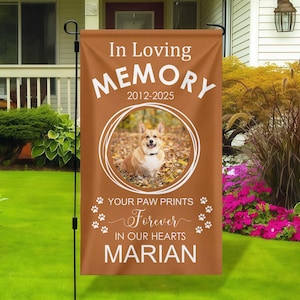 May include: A brown garden flag with the text "In Loving Memory" and "2012-2025". It features a photo of a corgi dog inside a white circle, with the words "Your Paw Prints Forever In Our Hearts" and the name "Marian".