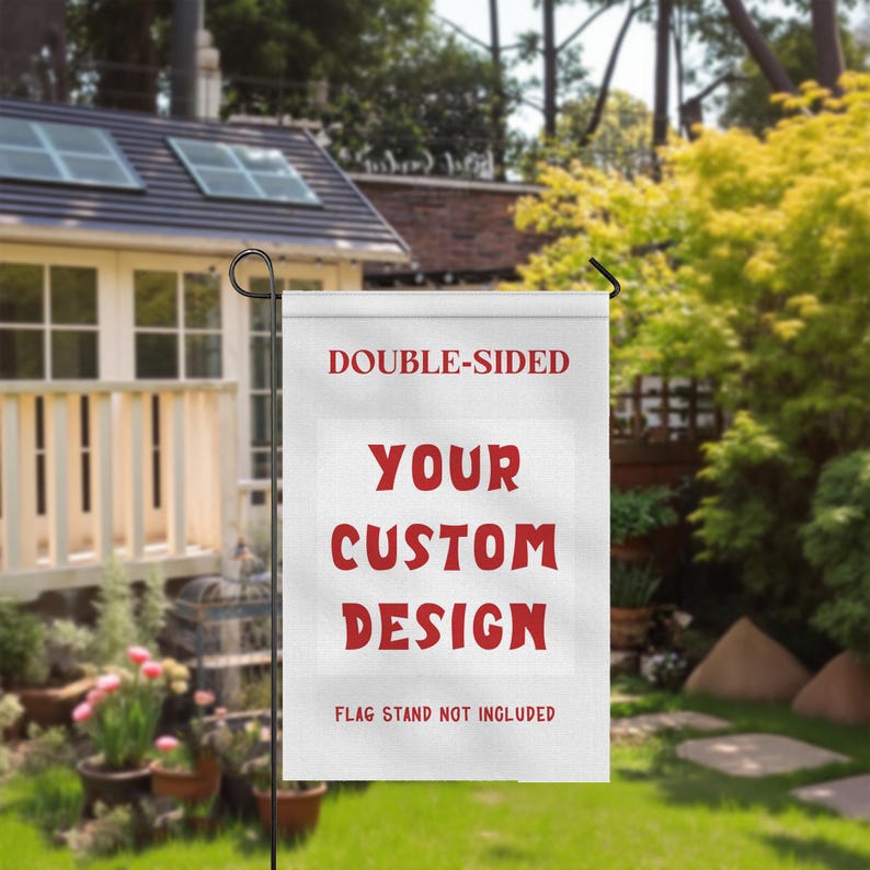 May include: A white, double-sided garden flag with the text "YOUR CUSTOM DESIGN" in red. The flag is displayed outdoors, with a house and garden in the background. The text "FLAG STAND NOT INCLUDED" is also visible.