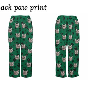 Op de afbeelding: Groene pyjamabroek met een zwarte pootprint en een kattengezichtpatroon. De tekst "Black paw print" staat bovenaan de afbeelding.