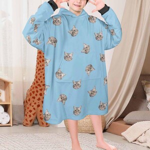 Puede incluir: Una sudadera con capucha azul claro con un estampado de gato por todas partes. La sudadera tiene un bolsillo grande en la parte delantera y est&aacute; hecha de un material suave y c&oacute;modo.