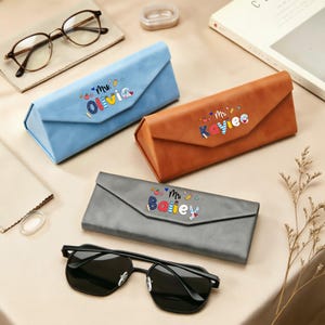May include: Three triangular folding glasses cases in light blue, tan, and gray. Each case features a personalized name printed on the front: Mrs. Olivia, Mrs. Kaylee, and Mrs. Bailey, accompanied by small colorful icons like apples and pencils.