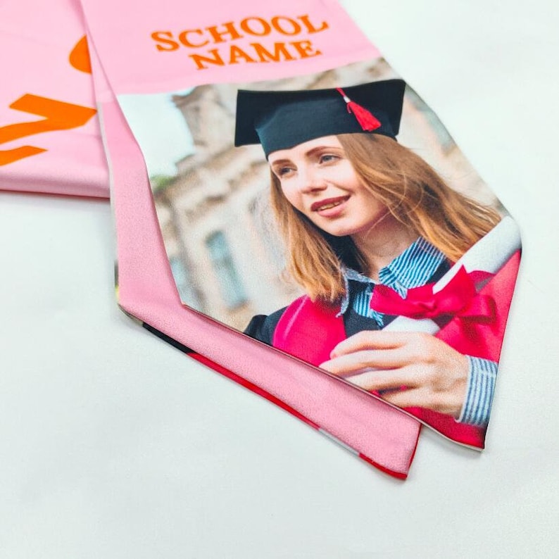 May include: Pink graduation sash with the text "SCHOOL NAME" printed in orange. The sash features a photo of a graduate wearing a black cap and gown, holding a diploma.