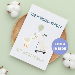 May include: A greeting card with the text "THE HORRORS PERSIST BUT WE STAY SILLY" and illustrations of geese. One goose wears a party hat and the other rides a scooter. A balloon is also featured. The card is on a wooden plate.
