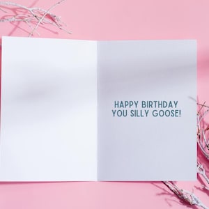 May include: Open white birthday card with the text "HAPPY BIRTHDAY YOU SILLY GOOSE!" in teal. The card is open on a pink background with white branches in the corners.