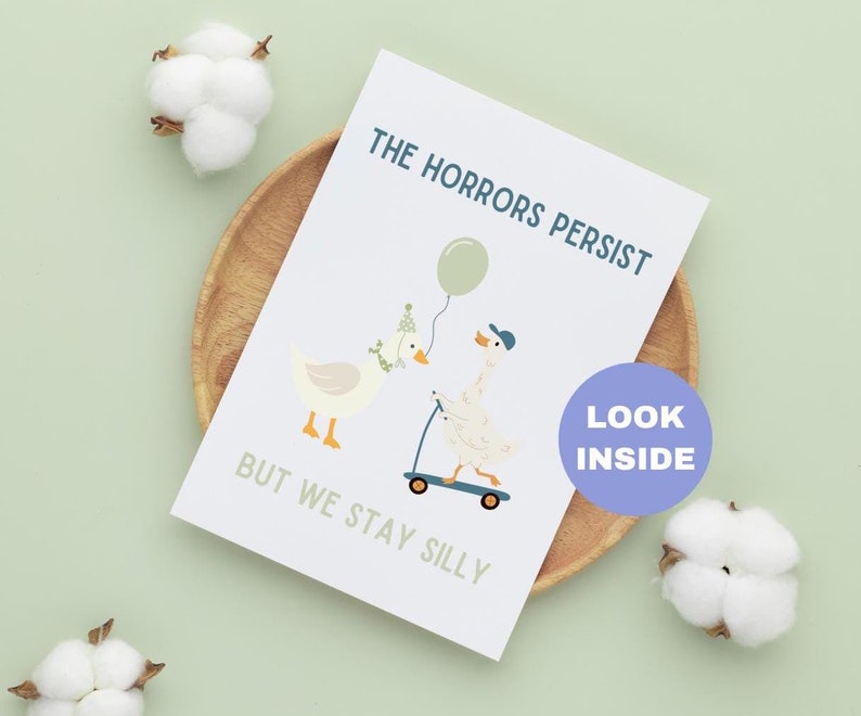 May include: A greeting card with the text "THE HORRORS PERSIST BUT WE STAY SILLY" and illustrations of geese. One goose wears a party hat and the other rides a scooter. A balloon is also featured. The card is on a wooden plate.
