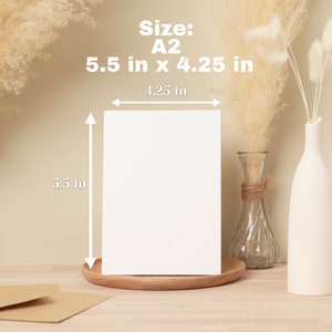 May include: A white card mockup on a wooden surface with a size of 5.5 inches by 4.25 inches. The card is sitting on a wooden coaster. There are two vases with dried pampas grass in the background.