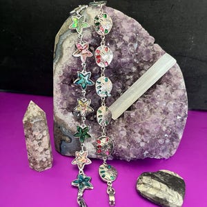 May include: Two silver bracelets with colorful star and circle charms. The bracelets are displayed on a purple background with a large amethyst geode and two smaller stones.