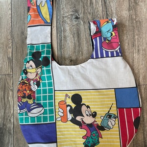 May include: A white canvas tote bag with a colorful Mickey Mouse design. The bag features Mickey Mouse in different poses, including holding a drink and a walkie-talkie. The bag has a round shape and two straps.