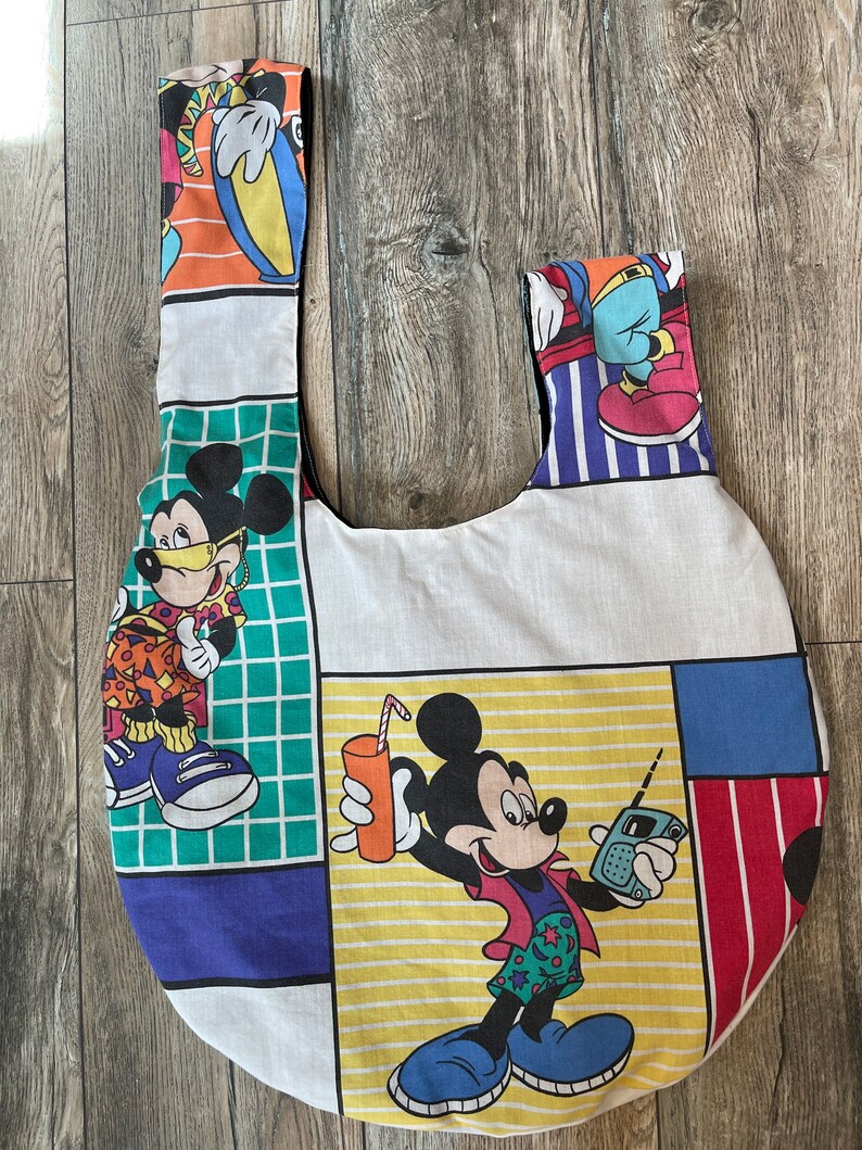 May include: A white canvas tote bag with a colorful Mickey Mouse design. The bag features Mickey Mouse in different poses, including holding a drink and a walkie-talkie. The bag has a round shape and two straps.