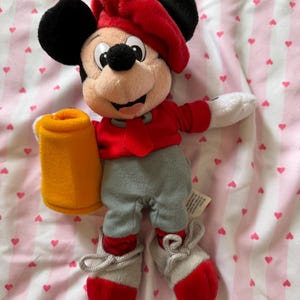 May include: A Mickey Mouse plush toy wearing a red beret, jacket, and shoes, with gray pants. The plush holds a yellow mug. The toy has black ears and a smiling face. The background is a pink and white striped fabric with heart shapes.