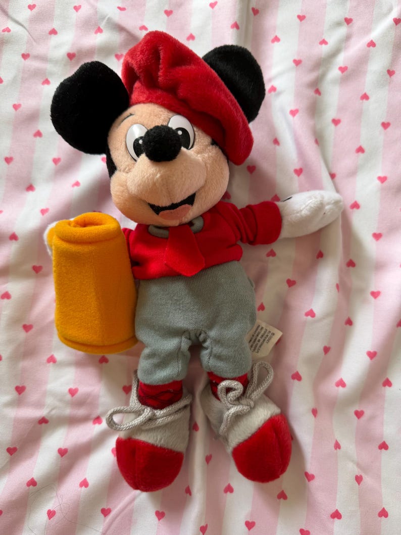 May include: A Mickey Mouse plush toy wearing a red beret, jacket, and shoes, with gray pants. The plush holds a yellow mug. The toy has black ears and a smiling face. The background is a pink and white striped fabric with heart shapes.
