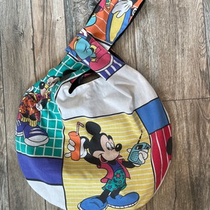 May include: A round white canvas tote bag with a black strap. The bag features a colorful patchwork design with Mickey Mouse and other Disney characters.