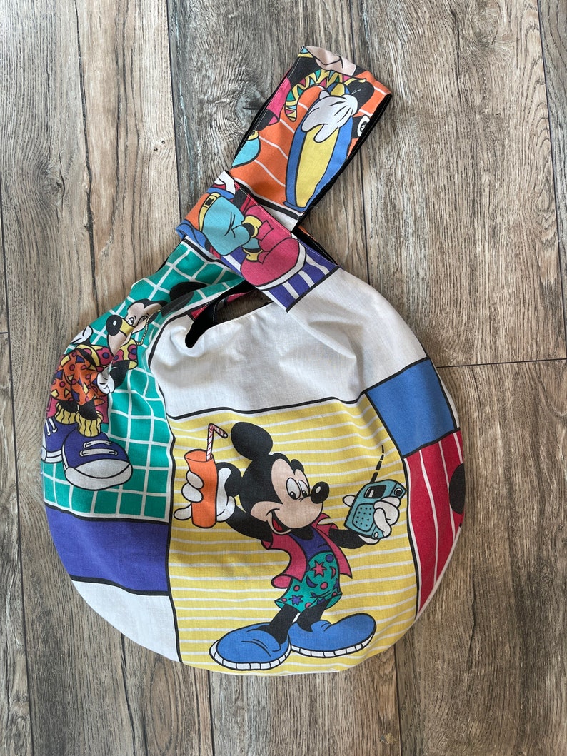 May include: A round white canvas tote bag with a black strap. The bag features a colorful patchwork design with Mickey Mouse and other Disney characters.