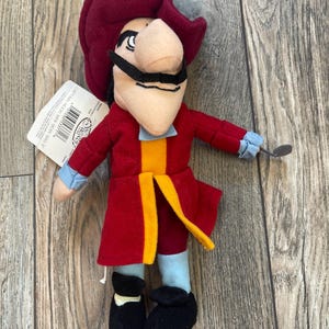 May include: A plush Captain Hook doll with a red coat, hat, and black mustache. The doll has a large nose, a yellow and orange vest, and a small metal hook. The doll is a Disney product.