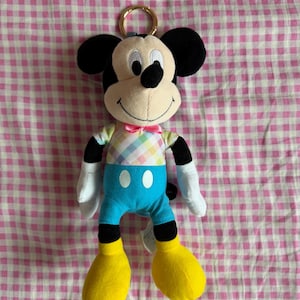 May include: A Mickey Mouse plush toy with a gold-colored keyring. The plush has a cream-colored face, black ears, and a black nose. It wears a pastel plaid shirt, blue shorts, yellow shoes, and a pink bow tie. The background is a pink and white checkered pattern.