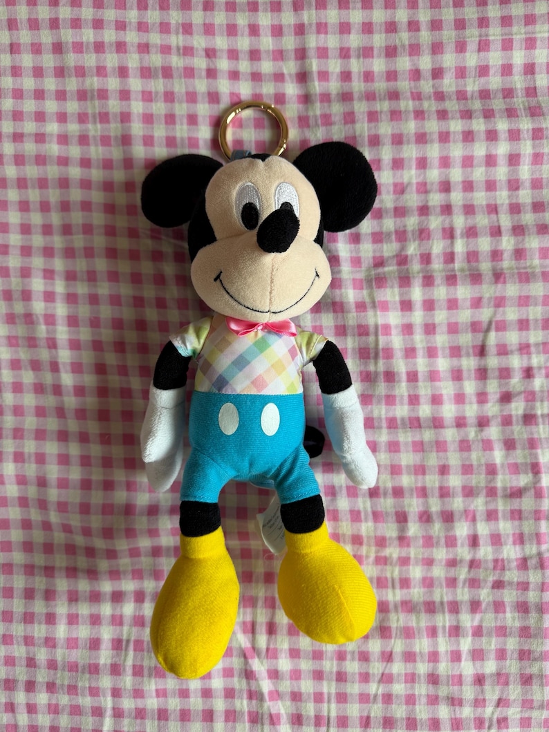 May include: A Mickey Mouse plush toy with a gold-colored keyring. The plush has a cream-colored face, black ears, and a black nose. It wears a pastel plaid shirt, blue shorts, yellow shoes, and a pink bow tie. The background is a pink and white checkered pattern.