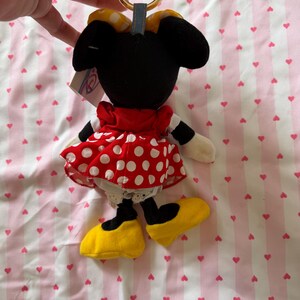 May include: A Minnie Mouse plush keychain. The plush has a black head and ears, a red dress with white polka dots, and yellow shoes. The keychain has a gold ring and a blue fabric loop.