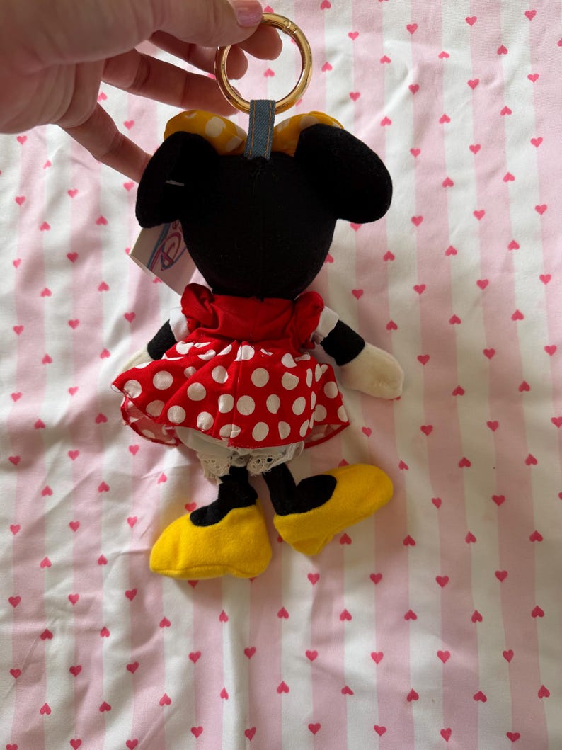 May include: A Minnie Mouse plush keychain. The plush has a black head and ears, a red dress with white polka dots, and yellow shoes. The keychain has a gold ring and a blue fabric loop.