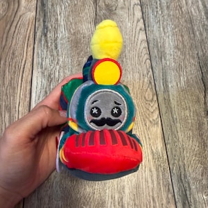 May include: A plush toy train with a smiling face, mustache, and star-shaped eyes. The train features a red front, a teal and green body, and a yellow smokestack. The toy is designed for children.