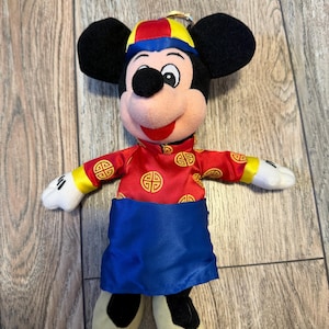 Pu&ograve; includere: Un peluche di Topolino con un abito rosso e oro in stile cinese, con una fascia blu e un cappello abbinato. Il peluche ha orecchie nere, viso rosa e scarpe color crema. L'abito presenta simboli dorati.