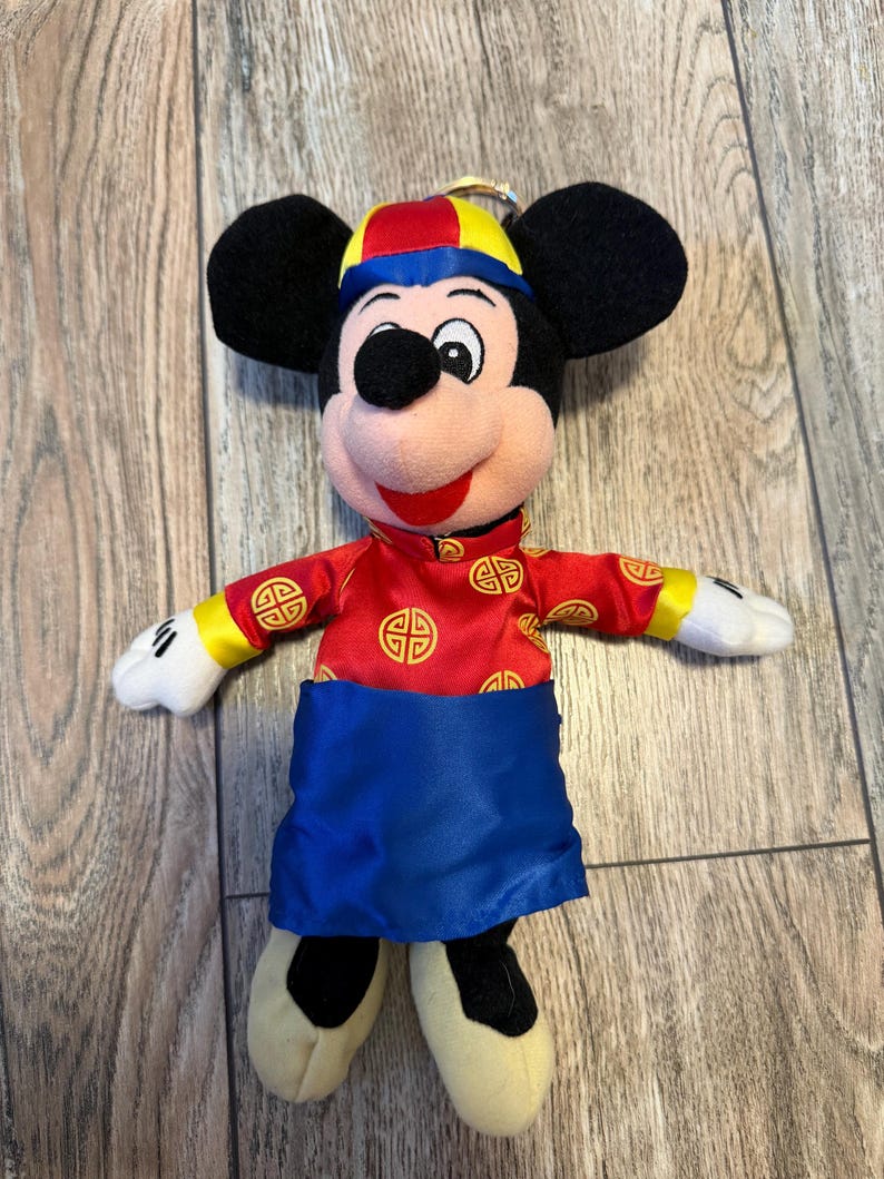 Pu&ograve; includere: Un peluche di Topolino con un abito rosso e oro in stile cinese, con una fascia blu e un cappello abbinato. Il peluche ha orecchie nere, viso rosa e scarpe color crema. L'abito presenta simboli dorati.