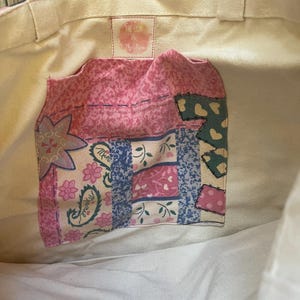 May include: A white canvas tote bag with a pink and blue patchwork fabric pocket. The pocket has a floral design and the text "The Lion & the Bird" is printed on the bag.