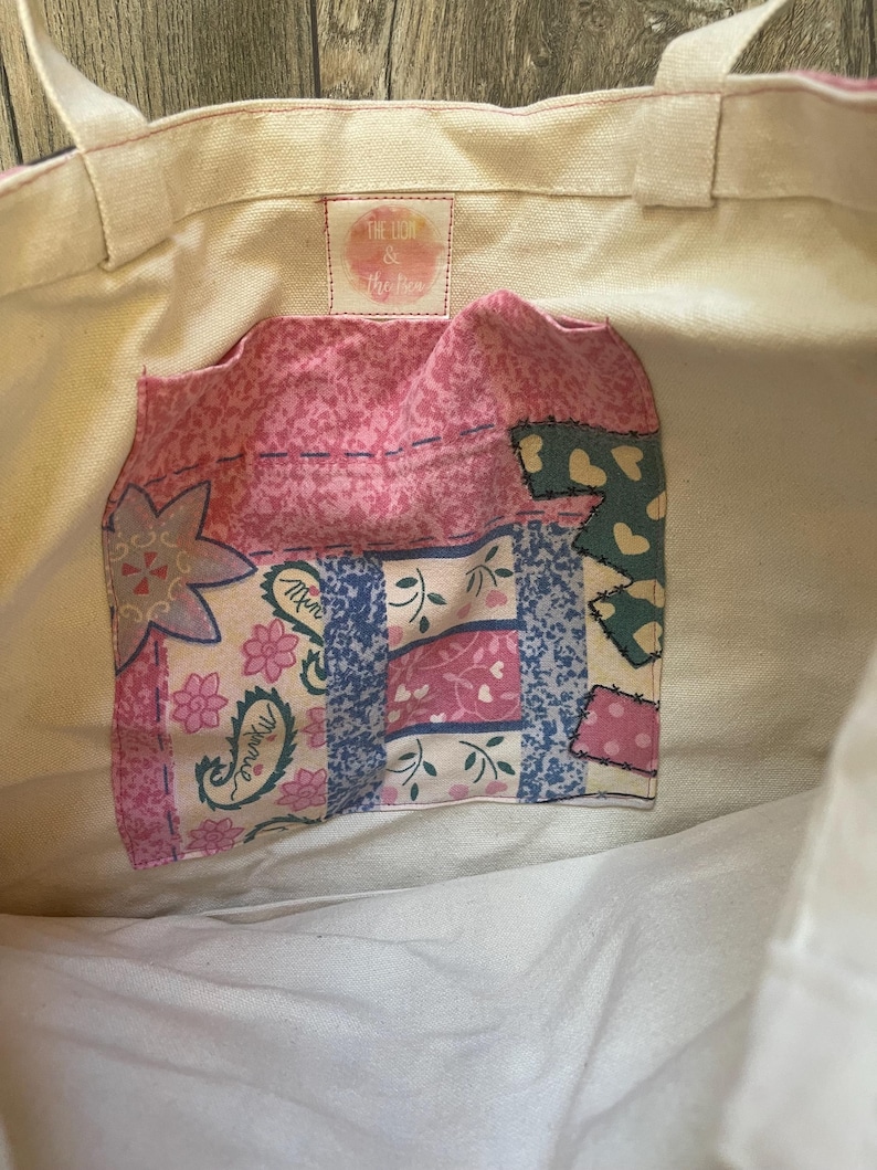 May include: A white canvas tote bag with a pink and blue patchwork fabric pocket. The pocket has a floral design and the text "The Lion & the Bird" is printed on the bag.
