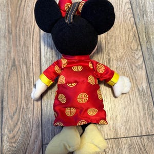 May include: A plush Minnie Mouse doll wearing a red dress with gold accents and a matching bow. The doll has black ears, white gloves, and yellow shoes. The dress features a repeating gold pattern.