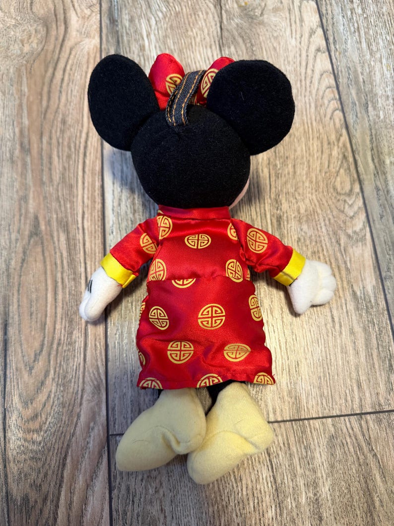 May include: A plush Minnie Mouse doll wearing a red dress with gold accents and a matching bow. The doll has black ears, white gloves, and yellow shoes. The dress features a repeating gold pattern.