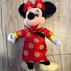 May include: A Minnie Mouse plush keychain wearing a red dress with gold accents and a matching bow. The dress features a repeating gold pattern. The plush has black ears, a pink face, and yellow shoes. A gold-colored keyring is attached to the top.