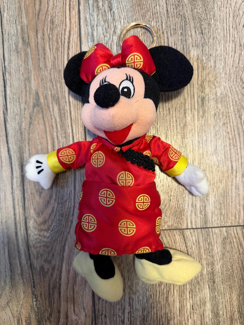 May include: A Minnie Mouse plush keychain wearing a red dress with gold accents and a matching bow. The dress features a repeating gold pattern. The plush has black ears, a pink face, and yellow shoes. A gold-colored keyring is attached to the top.