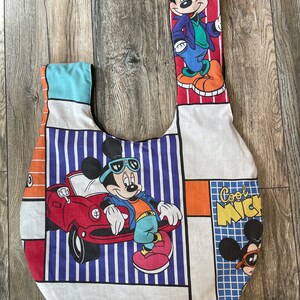 May include: A white fabric tote bag with a patchwork design featuring Mickey Mouse in different poses. The bag has a blue and white striped section, a red and orange section, and a blue and white checkered section. The bag also features the text "Cool Mickey".