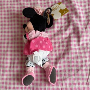 May include: A Minnie Mouse plush toy wearing a pink polka dot dress, black ears, and a pink bow. The toy has a gold-coloured ring attached to the head. The background is a pink and white checked pattern.