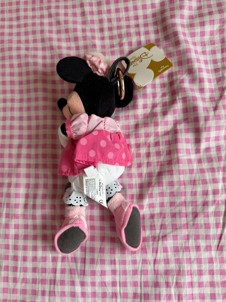 May include: A Minnie Mouse plush toy wearing a pink polka dot dress, black ears, and a pink bow. The toy has a gold-coloured ring attached to the head. The background is a pink and white checked pattern.