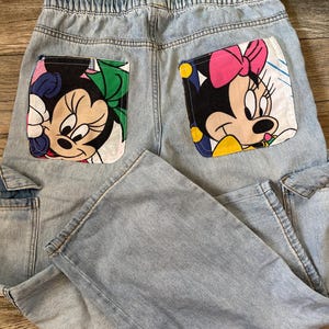 May include: Light blue denim pants with two back pockets featuring Minnie Mouse. One pocket has Minnie with a green bow, and the other has Minnie with a pink bow. The pants have an elastic waistband and cargo pockets.