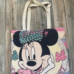 May include: A beige canvas tote bag with a patchwork design featuring Minnie Mouse. The bag has a large image of Minnie Mouse with a pink bow and the text "Simply the Best".