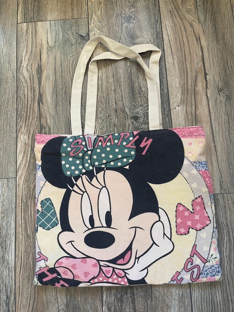 May include: A beige canvas tote bag with a patchwork design featuring Minnie Mouse. The bag has a large image of Minnie Mouse with a pink bow and the text "Simply the Best".