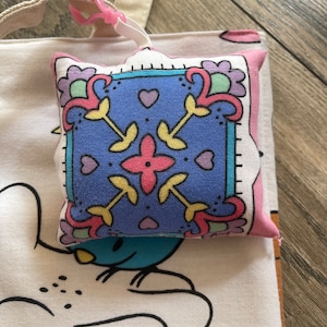May include: A small, square, decorative pillow with a blue center featuring a floral design in pink, yellow, and green. The pillow is attached to a white tote bag with a cartoon character design via a pink clip.