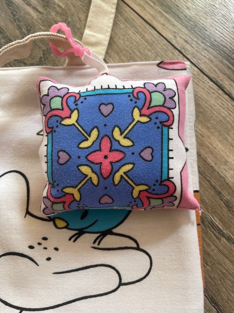 May include: A small, square, decorative pillow with a blue center featuring a floral design in pink, yellow, and green. The pillow is attached to a white tote bag with a cartoon character design via a pink clip.