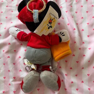 May include: A Mickey Mouse plush toy wearing a red jacket, gray pants, and red and gray shoes. The toy has black ears, white gloves, and a red hat with a gold ring. A Disney World tag is attached.