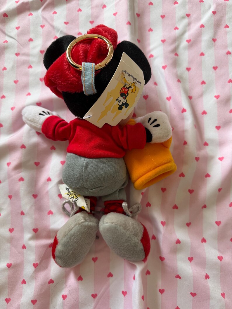 May include: A Mickey Mouse plush toy wearing a red jacket, gray pants, and red and gray shoes. The toy has black ears, white gloves, and a red hat with a gold ring. A Disney World tag is attached.