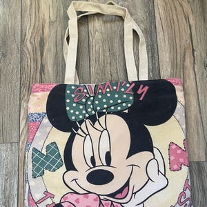 May include: A beige canvas tote bag with a colorful patchwork design featuring Minnie Mouse. The design includes hearts, polka dots, and the text "Simply the Best".