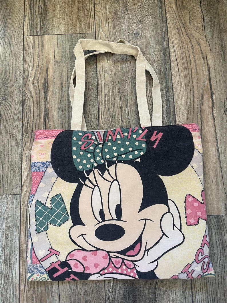 May include: A beige canvas tote bag with a colorful patchwork design featuring Minnie Mouse. The design includes hearts, polka dots, and the text "Simply the Best".