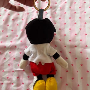 Op de afbeelding: Een Mickey Mouse pluche sleutelhanger. De figuur heeft een zwart hoofd, een wit shirt, een rode korte broek en gele schoenen. De sleutelhanger heeft een gouden ring en een blauwe stoffen lus.