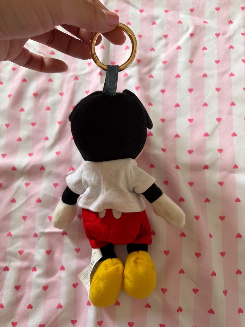 Op de afbeelding: Een Mickey Mouse pluche sleutelhanger. De figuur heeft een zwart hoofd, een wit shirt, een rode korte broek en gele schoenen. De sleutelhanger heeft een gouden ring en een blauwe stoffen lus.