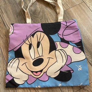 May include: A light blue canvas tote bag featuring a purple and white Minnie Mouse design. The bag has white canvas handles and the text "&copy; Disney" is printed on the bottom.
